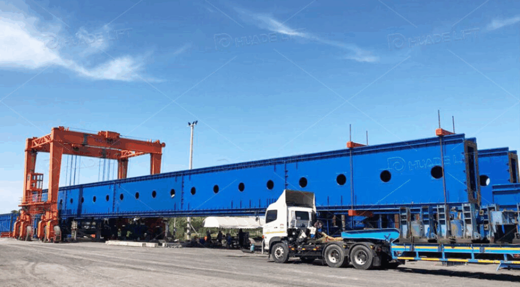 Double girder rubber tyred gantry crane lifting a long steel structure component at an industrial yard.